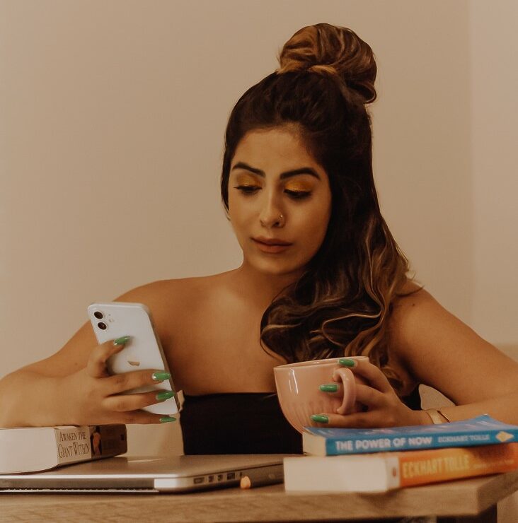 Marzia on her worktable. looking at her phone. There's a pink coffee mug! Books by Tony Robbins and Eckhart tolle.
