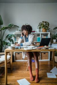 A woman organizing her desk while talking on the phone, surrounded by books and plants, symbolizing the importance of decluttering and maintaining an organized workspace for productivity.