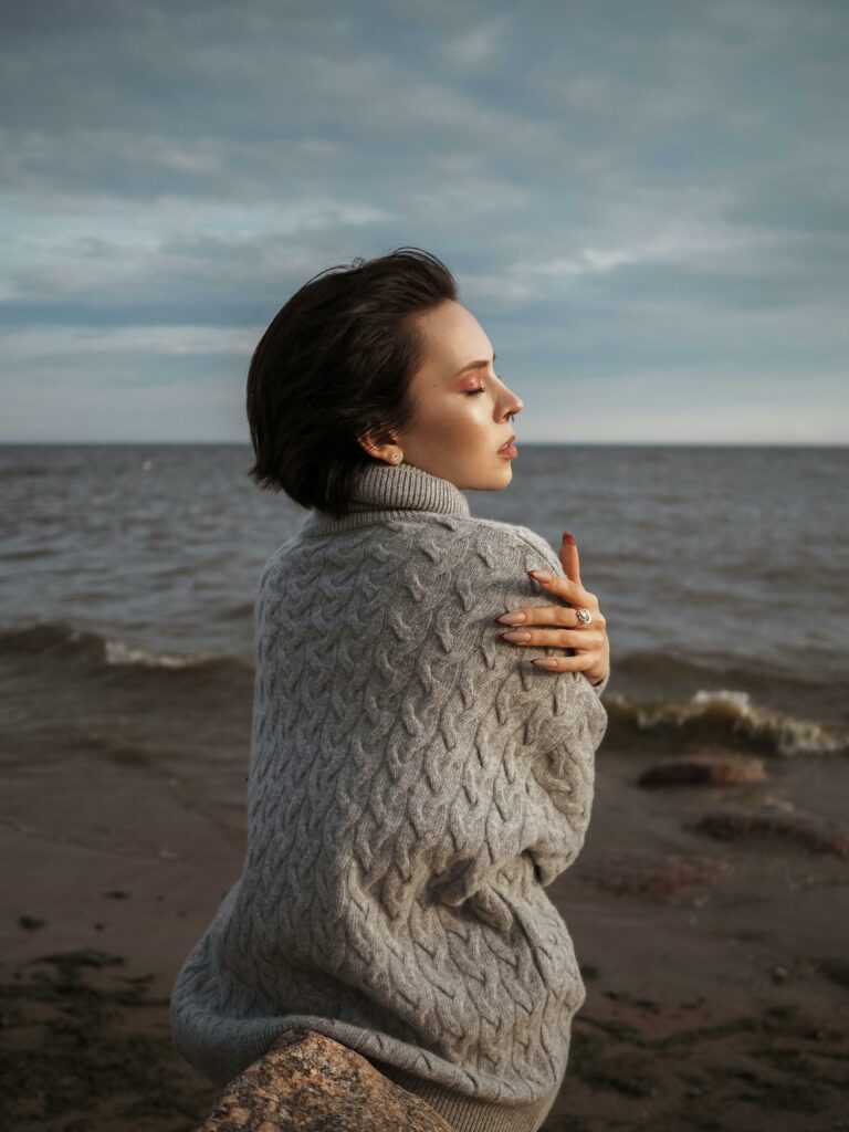 Woman sitting by the sea with eyes closed, embracing herself.